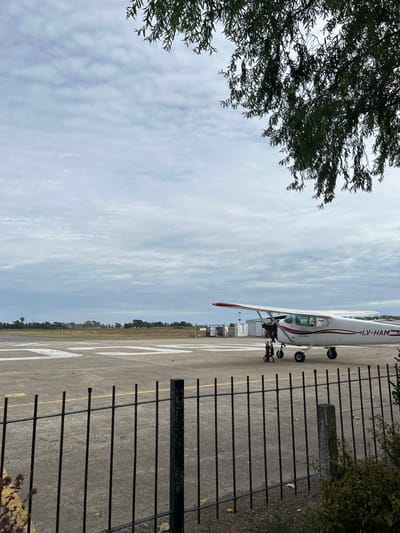 The image depicts an airfield under an overcast sky, viewed through a dark metal fence and partial tree foliage. The scene is located at an aerodrome in Chascomús, Argentina.

In the foreground, a black metal fence with vertical bars and horizontal rails spans the lower width of the frame. Green and brown shrubbery partially obscures the fence on the lower right and a yellow object is partially visible on the far left. Overhanging dark green tree foliage, with slender leaves, frames the top right corner of the sky.

The midground features a concrete or asphalt apron with white painted markings, including a prominent "30" numeral. Parked on this surface is a single-engine, high-wing propeller aircraft, predominantly white with red accents on the wingtips and tail. The aircraft bears the registration "LV-HAM" on its fuselage. The engine cowling of the aircraft is open or removed, exposing internal components, and a red portable jack stand is positioned beneath the nose/engine section. A smaller red object is on the ground near the front landing gear.

In the background, a line of dense green trees and distant low-lying vegetation extends across the horizon. Several small, white structures with black details, possibly airfield utility buildings or equipment, are visible to the right of the aircraft. The sky is uniformly cloudy with a light grey tone, indicating an overcast condition. No individuals are visible in the frame.