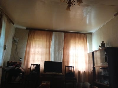 The image depicts a dimly lit living room in a home. The scene is dominated by a large window covered with sheer, patterned curtains, allowing diffused light to enter, suggesting it might be daytime. There is a television set on a wooden cabinet in front of the window, flanked by two wooden chairs. To the right, a tall glass-fronted cabinet displays various glass items on its shelves. On the left side of the room, a stack of items, possibly blankets or clothing, is piled on a surface. A ceiling light fixture with multiple lamps is visible at the top of the frame. The overall impression is one of a lived-in, perhaps slightly dated, interior. No people are visible in the image. The location context is Круглое, Russia.