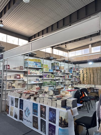 This image presents an indoor scene depicting a book exhibition or fair, located in Al-Ghizlaniya, Syria. The environment is a large hall with a high, corrugated metal ceiling from which industrial pendant lights and rectangular fixtures are suspended. Natural light is admitted through horizontal windows positioned high on the far wall.

In the foreground, a long table covered with a white cloth is laden with numerous stacks of books. Attached to the front of this table is a white banner displaying a logo resembling two stylized keys or pages, accompanied by the Arabic text "دار الرموز العربية" (Dar Al-Rumooz Al-Arabiya). Below the text, a grid of book covers is visible. Extending into the background are multiple metal shelving units, densely packed with thousands of books, arranged both horizontally in stacks and vertically on shelves. Behind the main foreground table, a smaller table holds a water bottle and a small white floral arrangement.

To the right of the scene, several black plastic chairs are visible, one of which has a black jacket or coat draped over it. A decorative gold-colored screen featuring geometric patterns partially obscures the background on the far right. The floor throughout the visible area is covered with grey carpeting. A partial view of a person's arm and side is visible on the extreme right edge of the frame. No direct actions or interactions are clearly depicted, but the arrangement of books and furniture indicates a retail or display setting.