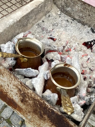 Two cezves (Turkish coffee pots), each containing coffee, are nestled among white ash and glowing embers within a metal fire pit. The pit sits on a paved surface, likely in an outdoor setting. The overall location is speculated to be Konya, Turkey.