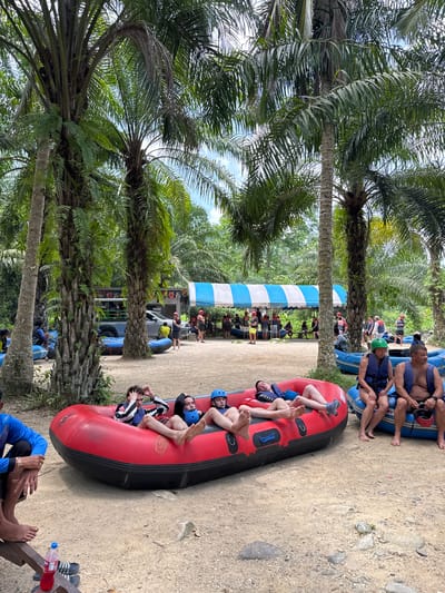 The image shows a scene in Tambon Song Phraek, Thailand. Several rafts are present on a dirt or sandy surface, with people relaxing in them. Some individuals are wearing helmets and life jackets, suggesting a water activity like rafting. Other people are standing and sitting around the rafts. Palm trees are prominent in the foreground and background. A blue and white striped structure is also visible in the background. The environment appears to be a recreational area near a river or body of water.