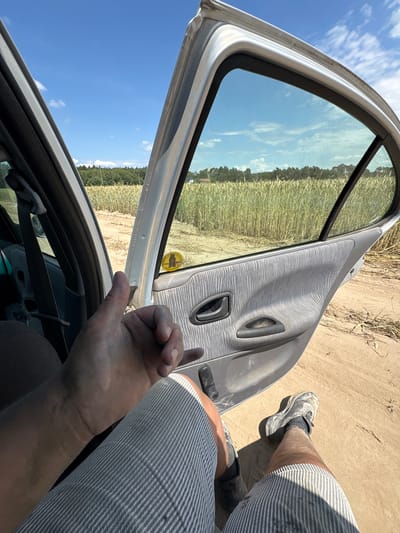 The image depicts a person's hand giving a thumbs-up gesture inside a car with the doors open. The car is parked on a dirt road next to a field of wheat under a clear blue sky with scattered clouds. The interior of the car shows a grey fabric door panel with a black door handle and a yellow sticker. The person is wearing striped shorts and dirty athletic shoes. The background features a dense forest line. The exact location in Opatów, Poland cannot be determined from the image.