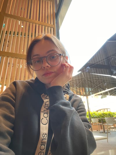 A woman wearing glasses and a Calvin Klein sweatshirt is sitting in an indoor space, likely a café or restaurant, in Marcianise, Italy. The interior has wooden slatted walls and the seating area has chairs. The image is taken from a slightly elevated angle, looking down at the woman. Outside the window, there is foliage and architectural features.