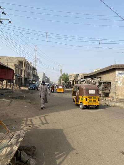 The image depicts a street scene in Kofar Mazugal, Nigeria, under a clear, light blue sky. A paved asphalt road, with unpaved dirt shoulders, extends into the distance.
In the foreground, a male figure walks on the left dirt shoulder, facing towards the viewer. He wears a light-colored, long robe and a blue face mask. To the immediate left, a makeshift wooden structure rests on large rocks.
Vehicles populate the road. In the midground-right, a yellow three-wheeled auto-rickshaw with a brown canopy drives away from the viewer, displaying "Keke MBA" on its rear. Further down the road, another yellow auto-rickshaw and a red motorcycle are also moving away. On the left shoulder, a dark-colored sedan and a white van are parked.
Buildings line both sides of the street. On the left, multi-story concrete structures, some appearing incomplete with exposed rebar, rise behind the parked vehicles. In the distant background-left, a red and white lattice tower is visible. On the right side, a row of single-story buildings with rough, unpainted facades and corrugated roofs extends into the background. One building on the right has a partial sign reading "ZUBAIRU E...". Numerous overhead power lines traverse the sky, supported by utility poles. Shadows on the ground suggest strong sunlight.