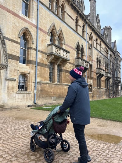 An adult male is observed from a right-rear perspective, pushing a dark-framed stroller with an olive-green fabric component. The man wears a dark blue winter coat, dark trousers, dark athletic shoes, and a navy blue knit bobble hat with "LONDON" written in white and red lettering, topped with a red/pink pompom. A small child is partially visible within the stroller. A dark brown quilted bag and a light-colored plush toy are attached to the stroller handle.
The scene is set outdoors on a paved path constructed from irregular, light-colored cobblestones, which appears damp with a visible puddle towards the right. To the right of the path lies a strip of green grass. In the background, a large, multi-story building of the Oxford University Museum of Natural History in Oxford, United Kingdom, dominates the view. The building exhibits Victorian Gothic architecture, constructed from light yellow stone with distinct horizontal reddish-brown banding. It features numerous arched windows, some with decorative iron grates, and multiple small, ornate stone balconies. The upper levels include prominent gables, chimneys, and turret-like structures. Sections of the building, particularly towards the right, are partially covered in dark green ivy. The sky is overcast and light grey, providing diffused natural light.