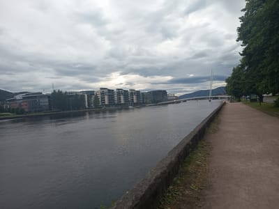 The image shows a wide, calm river flowing through a city under a cloudy sky. On the left bank, there are modern apartment buildings and office blocks. On the right bank, a paved pathway runs along the river, bordered by trees and grass. In the distance, a distinctive white suspension bridge spans the river, with its tall, slender pylons reaching towards the sky. A few people can be seen walking on the pathway. The overall impression is of an urban waterfront with a mix of residential and commercial architecture, and recreational space. This scene is likely the Drammenselva river in Drammen, Norway, given the distinctive bridge.