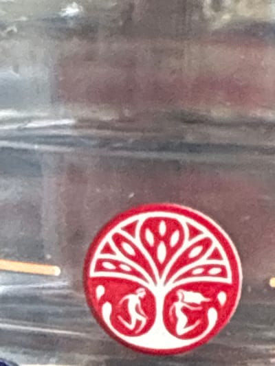 The image shows a red and white circular decal with a stylized tree design. The tree has a trunk that splits into two, with two figures, one on each side, reaching upwards. The branches of the tree are depicted as flowing, organic shapes. The decal is placed on a dark, possibly metallic or glass surface, with horizontal lines suggesting it is on a container or a vehicle. There is a blurred background that appears to be a building or wall, with some horizontal elements that could be windows or shelves. The lighting is diffused, creating a somewhat muted overall tone. The location of this decal in Bad Hersfeld, Germany, is not discernible from the image itself.