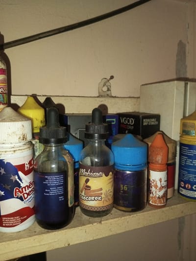 A cluttered shelf displays an assortment of vaping liquid bottles. "American Dessert," a blue liquid in a bottle, and "Mahima Chocoroz" are visible, alongside bottles with labels like "VGOD Pro Drip," a nicotine level of "36 milligrams" and a note about not using during pregnancy. Indonesian text and ingredients like vegetable glycerin are featured on the labels. The shelf also contains a Rebuildable Drip Atomizer box. Dust and grime are apparent, adding to the chaotic feel of the storage space.