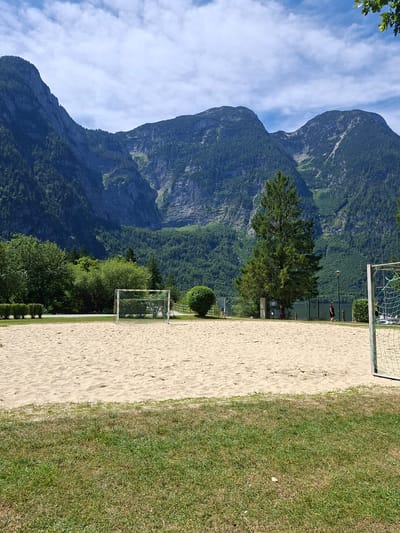 The image depicts a sand volleyball court in Obertraun, Austria, with a mountainous backdrop. The court is situated on a sandy surface with a grassy area in the foreground. Two soccer goals are visible on the court, one in the mid-ground and another partially visible on the right side of the frame. A tall evergreen tree stands behind the court, and dense green foliage covers the lower slopes of the imposing mountains that dominate the background. The sky is partly cloudy, with blue patches and white clouds scattered across it. A few people are present in the scene, with one individual walking along a path behind the court and another person visible near the soccer goal on the right. The overall scene conveys a sense of leisure and outdoor activity in a scenic natural setting.