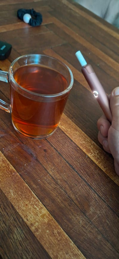 A glass mug filled with amber-colored liquid, likely tea, sits on a wooden table. To the right of the mug, a hand holds a rose gold colored electronic cigarette device, which is lit, emitting a faint glow from its indicator light. In the background, a black hair scrunchie with a white pearl-like accent lies on the table. The table surface exhibits a wood-grain pattern, with distinct planks arranged diagonally. The scene appears to be indoors, possibly in a home in Shakhtarsk, Ukraine.
