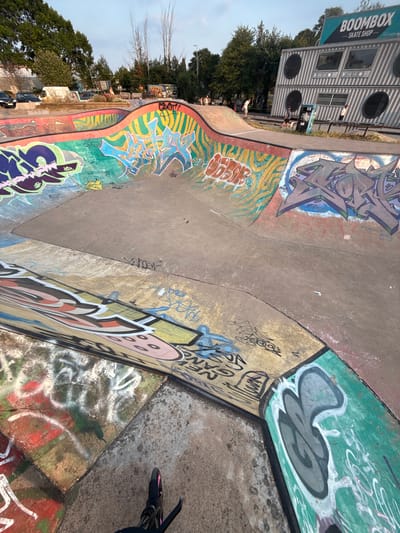 The image shows a graffiti-covered concrete skatepark on a sunny day. In the background, a building designed to resemble a boombox with the text "BOOMBOX SKATE SHOP" is visible. Several people are present in the skatepark, some of whom appear to be skateboarding or scootering. The skatepark features ramps and bowls, all adorned with colorful graffiti art. The overall environment suggests a recreational area in an urban setting, likely Hereford, United Kingdom, as indicated by the presence of the "Boombox Skate Shop" which is a known location in Hereford. The lighting suggests it is late afternoon.