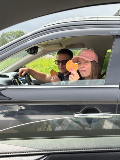 Two people are inside a dark-colored SUV, possibly a Mitsubishi Outlander, with the windows rolled down. A male driver wearing glasses and a black t-shirt holds the steering wheel with his left hand and a round waffle cookie in his right hand. A female passenger, wearing a pink baseball cap with "Atelier Paris" embroidered on it, is on the right side of the car. She is holding a similar waffle cookie with her right hand and looking at the camera. Both individuals appear to be of young adult age. The interior of the car is visible, including the dashboard and car door. Outside the car, green trees and foliage are seen under an overcast sky. The scene is consistent with being in a vehicle in a rural or suburban setting, possibly on a road trip, and the location of Теплоозёрск, Russia, is a small settlement known for its natural surroundings, which aligns with the visual evidence.