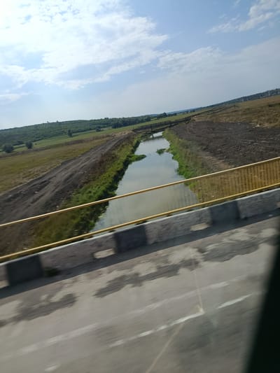 This image captures a landscape scene in Țipala, Moldova, viewed from a bridge. In the foreground, the surface of a road, likely asphalt, is visible with indistinct markings and shadows, suggesting motion blur from a vehicle. A concrete barrier with a yellow metal railing runs along the edge of the bridge. Beyond the bridge, a narrow waterway, possibly a canal or a small river, flows through the terrain. Its banks are lined with green vegetation and bare earth or disturbed soil on one side, indicating some form of construction or land management. The waterway is crossed by a simple wooden bridge in the mid-ground. The background consists of gently rolling hills covered with green fields and scattered trees. The sky is partly cloudy with a bright, clear blue expanse.