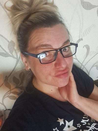 A close-up, chest-up selfie portrays a female subject indoors in the city of Yegoryevsk, Russia. The individual, appearing middle-aged, has light-colored hair gathered in a high, messy bun. She is wearing black-framed glasses, with visible reflections in the lenses, and is looking directly at the camera. Her lips are slightly pursed in a pouting expression. Her right hand (viewer's left) is placed against her jawline. Accessories include a thin gold chain necklace and a small gold hoop earring in her visible left ear. She wears a dark-colored, short-sleeved t-shirt adorned with white star patterns and partial text. The background is a light-colored wall with a subtle, repeating pattern of dark, stylized floral or leaf motifs.