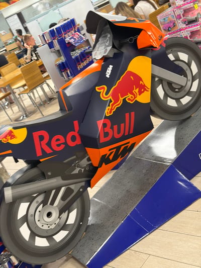 The image shows a life-size cardboard cutout of a KTM Red Bull racing motorcycle, displayed indoors, likely in a retail environment. The motorcycle is predominantly navy blue with prominent Red Bull branding, including the red bull logo and the text "Red Bull" in large red lettering. Orange accents are visible on the fairings and other parts of the motorcycle. The wheels are black with white spokes. In the background, there are shelves stocked with Red Bull cans and other products, suggesting a point-of-sale display. Several people are visible in the background, some appearing to be customers or staff. The flooring appears to be a light-colored wood or laminate. The setting resembles a convenience store or a supermarket aisle. There is no indication of the city of Due Carrare, Italy.