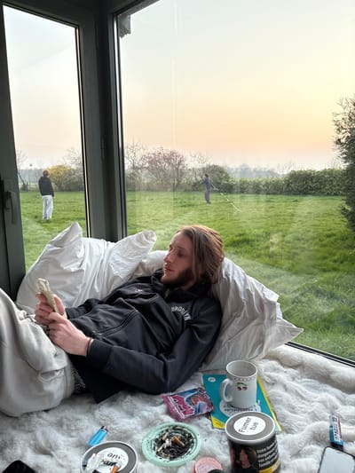 The image displays an indoor and outdoor scene at Bû, France, during what appears to be either sunrise or sunset, indicated by the soft orange-yellow gradient in the sky. Inside, a male individual, estimated to be in his late teens or early twenties with long brown hair, a beard, and a mustache, reclines against white pillows on a fluffy white rug adjacent to a large window. He wears a black zippered hoodie with "BROOKLYN" inscribed on the chest and light-colored pants. Both hands are actively engaged with a light-colored smartphone. On the rug beside him are a blue disposable lighter, a circular green silicone ashtray containing multiple cigarette butts and ash, a metal tin displaying "Fumer tue" (Smoking kills) and French warning labels, a white ceramic mug with red cherry decals, a yellow book or magazine, and a colorful snack wrapper. Through the large window, a spacious green lawn is visible, bordered by trees and bushes. In the left-midground of the outdoor scene, a person in a dark jacket and light pants stands near a white spherical object on the grass. Further to the right, a second individual in dark clothing holds a long, thin object, with a white line extending across the lawn from their approximate location.