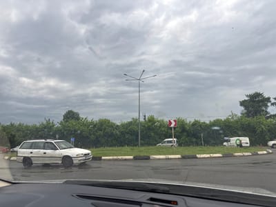 The image captures a cloudy day at a road intersection in Samtredia, Georgia. In the foreground, the dashboard of a vehicle is visible. To the left, a white station wagon is parked on the side of the road, facing right. Further down the road, a silver van is seen driving on the right side of the road. Behind the silver van, a white van with green circular logos is parked. A traffic sign with a red and white arrow indicating a sharp curve to the right is posted. The background is dominated by lush green trees and a grey sky. The road surface appears wet, suggesting recent rain.
