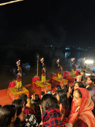 This is a nighttime photograph taken from an elevated perspective, overlooking a religious ceremony taking place by a body of water, likely the Tawi River in Jammu, India. The scene is illuminated by torches and the distant lights of the city.
In the foreground, a crowd of people, mostly women and children, are watching the ceremony. They are dressed in casual clothing, with some women wearing headscarves. Their faces show expressions of attention and engagement.
On a raised platform in the middle ground, several individuals dressed in traditional attire, consisting of maroon tunics and yellow lower garments, are performing a ritual. They are holding lit torches, creating a dramatic effect with the flames and smoke. Each performer stands on a decorated platform, adorned with flowers. Behind them, on the water, are two boats, one blue and one white, also with people on board.
The environment appears to be a riverbank or a ghat, with the water reflecting the lights. The background is dark, with the faint outline of buildings and lights indicating the presence of a city. The overall atmosphere suggests a cultural or spiritual event, possibly an Aarti ceremony, a common practice in India. The photograph captures a moment of collective experience and devotion.