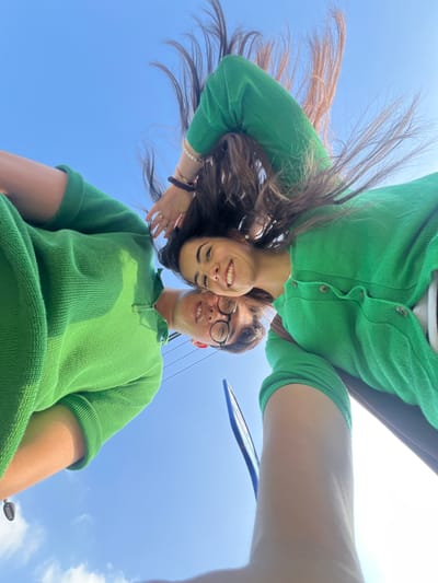 This media file is a low-angle, wide-shot selfie of two people against a bright blue sky with scattered clouds. The subjects, a woman and a man, are wearing matching green knit tops. The woman is on the left, her head tilted back with her long, dark hair flowing upwards and to the left, obscuring part of the sky. She has fair skin, is smiling broadly, and is wearing a pearl earring and several bracelets on her left wrist. The man is on the right, also smiling and wearing black-rimmed glasses. He has short dark hair and is wearing a green collared shirt with a half-zip. The perspective suggests they might be looking up from below, or the photo was taken while lying down. The overall mood is joyful and casual, possibly indicating a sunny day outdoors. The setting appears to be an open area, likely during daylight hours, given the bright sky and sunlight. There are no visible text elements in the image.