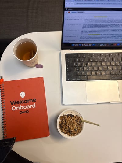 An overhead shot depicts a white table surface in Nes Ziona, Israel. A silver laptop, appearing to be a MacBook Pro with a Hebrew keyboard layout, occupies the right side, displaying an "elastic" Kibana dashboard on its screen. The screen shows data logs with timestamps (e.g., Oct 21, 2023), cURL commands, and mentions of "kibana.moovit.info" and "backend-services-app". To the left of the laptop, a white paper cup holds a light brown liquid, likely tea, with a purple-labeled tea bag (bearing Hebrew text "מרגנית", Marganit) submerged. Below the cup, a red spiral-bound notebook is positioned, displaying a white "Welcome Onboard" text with a smiling face logo above and two connected circles below. An orange pen is partially visible near the top left of the notebook. In the lower right quadrant, a white bowl contains a serving of yogurt topped with granola, and a light-colored spoon rests within the contents. A dark gray upholstered surface, possibly a couch or chair, is visible in the upper left background.