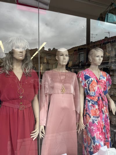 The image shows a shop window display featuring three mannequins dressed in women's clothing, with reflections of the sky and buildings visible on the glass. The mannequins are arranged from left to right.

The leftmost mannequin wears a pleated, short-sleeved red dress with a matching belt and a gold necklace featuring a star pendant. It has a gray, wavy wig.

The central mannequin is dressed in a light pink, sheer overlay dress with long sleeves. It has a bald head.

The rightmost mannequin wears a vibrant, floral-print dress with short, ruffled sleeves. The dress has a V-neck and is tied at the waist. It has short blonde hair and wears a gold pendant necklace.

The reflections in the window show a cloudy sky and the upper stories of buildings, suggesting an urban or suburban setting. The shop interior is visible behind the mannequins, with racks of clothing and accessories. One reflection shows a sign that appears to read "LA BAGUETTE DOBRE". Based on available information, this business is located at 26 Rue de la République, 77270 Villeparisis, France.