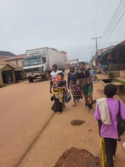 The image captures a street scene in Tangu, Nigeria, under an overcast sky. In the foreground, a young boy, seen from behind, walks away from the camera, wearing a purple t-shirt and black pants, with a white cloth draped over his shoulder and a dark backpack. To his left, three women are walking towards the left side of the frame. The woman closest to the foreground wears a colorful patterned dress and carries a black purse. The second woman from the foreground is in a blue top and a patterned skirt, carrying a blue bag. The third woman wears a patterned brown and black jacket over a blue top and a patterned skirt, and is carrying a bag and some rolled items. A white Mercedes-Benz truck is parked on the road, with its hazard lights on. The road surface is asphalt with visible cracks and some puddles. On the right side of the street, there are open market stalls with various goods displayed, including chairs and clothing. On the left side, there are buildings, including one with a sign that appears to read "INDO RAMA". Power lines are visible overhead. The overall scene suggests a bustling market environment.