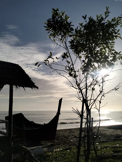 The image captures a beach scene in Sagoe, Indonesia. In the foreground, there's a silhouetted wooden boat nestled under a thatched roof structure. To the right of the boat, a tree with sparse leaves is silhouetted against the bright sunlight reflecting off the ocean. The beach stretches towards the calm water, and the sky is partly cloudy. There are no people visible in the scene. The image has a stark contrast, with the foreground appearing dark and the background, especially the ocean, brightly lit.