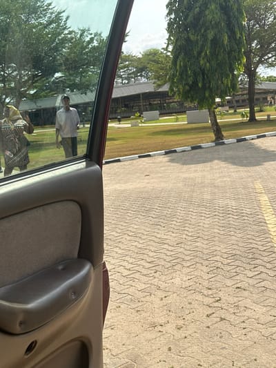 The image depicts an outdoor scene in Ozuaha, Nigeria, viewed from the interior of a vehicle, with a tan-colored door panel visible on the left side of the frame. The scene outside the vehicle features a light-colored, interlocking paver surface in the foreground, with a partial yellow line visible on the right edge. This paved area abuts a curb painted with alternating black and white stripes. Beyond the curb lies a maintained grassy area, interspersed with mature trees.
Two individuals are visible on the left side, appearing either reflected in the vehicle's window or seen through it. One individual, partially obscured, wears a patterned garment and a head covering, with an arm raised. The second individual, a male, is seen walking on the grass, dressed in a light-colored short-sleeved shirt and dark trousers. In the midground and background, multiple long, low-rise buildings with corrugated or metal roofs are visible, surrounded by additional trees and greenery under a bright sky.