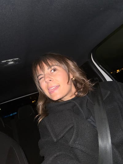 A woman with brown hair and bangs smiles slightly, wearing a black coat and seatbelt in what appears to be a car. The car's interior is dark, with a glimpse of city lights visible through the window. The woman has light skin, long eyelashes, and is wearing a gold earring. The angle suggests she may be looking up at the camera.