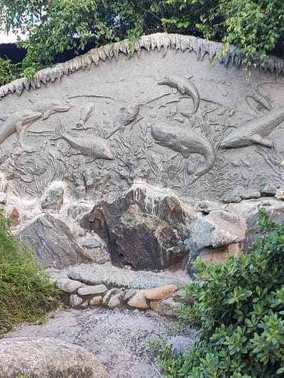 The image depicts a section of a bas-relief carving on a stone wall, likely part of an outdoor decorative feature in Bac Nha Trang Ward, Vietnam. The carving features marine life, including several fish and what appear to be dolphins or porpoises, depicted in various swimming positions. The relief is textured and appears to be made of grey stone, with some areas showing weathering. Below the carved section, there are natural rock formations of various sizes and colors, with some smaller, rounded stones arranged as a border in the foreground. Lush green foliage, consisting of trees and bushes, frames the top and right sides of the scene, suggesting an outdoor, possibly garden or park setting. The ground in the foreground is a mixture of gravel and small stones. There are no people visible in the image. The overall impression is of a man-made artistic element integrated into a naturalistic environment.