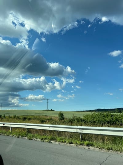 The image depicts a scenic landscape with a bright blue sky filled with a mix of fluffy white and darker, overcast clouds. In the foreground, a section of asphalt road is visible with a white metal guardrail separating it from a grassy embankment and fields beyond. The fields are a vibrant green, suggesting agricultural land, possibly wheat or barley. In the middle ground, several tall electricity pylons stand silhouetted against the horizon, indicating the presence of power lines. The background shows a gently rolling hill covered in trees and vegetation. The overall impression is one of a rural environment under a dynamic sky. The location is identified as Vakarel, Bulgaria. No people or specific actions are depicted.