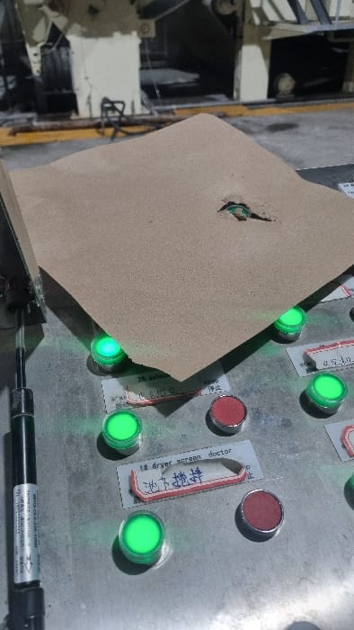 The image displays an industrial control panel, partially covered by a piece of light brown, roughly torn cardboard or paper. The panel is metallic and features an array of circular buttons or indicator lights. Many of these lights are illuminated green, suggesting an active status, while some are red and unlit.
Labels or stickers are affixed to the control panel next to some buttons. Visible text includes "1# dryer screen doctor" in English and Chinese characters "滤下料机" (Lǜ xiàliào jī, meaning "Filter discharge machine" or "Filter unloading machine") on one label. Another partially visible label near a red button shows the Chinese character "停止" (Tíngzhǐ, meaning "Stop"). The cardboard covers some of the buttons and labels.
In the lower right foreground, a black gas strut or damper is visible, bearing the text "0870-15-E-210-700N". The background, partially obscured, shows mechanical components of what appears to be industrial machinery, indicating an indoor factory or production setting. There are no people visible, and no specific activity is taking place beyond the apparent operational status of the control panel. The scene is illuminated by artificial lighting.
