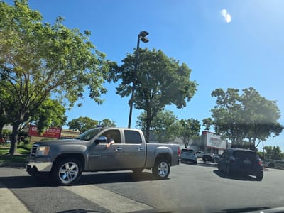 The image captures a sunny day scene in West Sacramento, USA, likely near a commercial area. In the foreground, a grey pickup truck is driving on a paved road, with a driver visible inside wearing sunglasses. The truck has chrome wheels and a short bed. Behind the truck, several other vehicles are present, including a dark-colored hatchback with a license plate starting with "7MT". Further in the background, a building with a red and white sign that reads "GROCERY OUTLET" is visible, surrounded by trees and a street light. The sky is clear and blue.