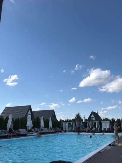 The image shows a contemporary resort pool area on a sunny day in Slobozhanske, Ukraine. A man stands by the edge of the clear blue water, while other people are seen relaxing on lounge chairs or swimming in the pool. In the background, modern, dark-roofed cabins and white gazebos are visible, surrounded by neatly trimmed trees. The sky is a bright blue with scattered white clouds. A young girl in a white swimsuit stands on the wooden deck of the pool, holding a hula hoop.