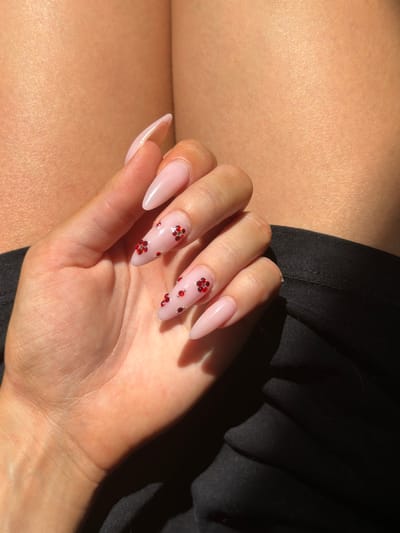 The image shows a close-up of a person's hand with stiletto-shaped, light pink acrylic nails, each adorned with small red rhinestones arranged in a floral pattern. The hand is positioned over a black, textured fabric, possibly clothing. The background consists of skin, likely of the same person, illuminated by strong sunlight casting shadows. The scene is indoors, as indicated by the quality of light and shadow, and there are no discernible elements that would place it specifically in Prosena, Bulgaria. The image is a still shot, focusing on the aesthetics of the manicure.