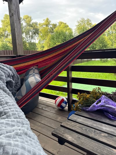 The image depicts a relaxed scene on a wooden deck, presumably in Gavshino, Russia. A colorful striped hammock is stretched across the frame, partially occupied by a person covered with a grey, textured blanket and a pillow with a blue and white geometric pattern. Next to the hammock, on the wooden floor, rests a red, white, and blue ball. To the right, a low wooden table holds a bunch of dried yellow and green herbs tied with a purple plastic bag. The background reveals a lush green lawn bordered by a dark wooden fence, with trees and a cloudy sky visible beyond. The overall impression is one of a quiet, outdoor leisure space.