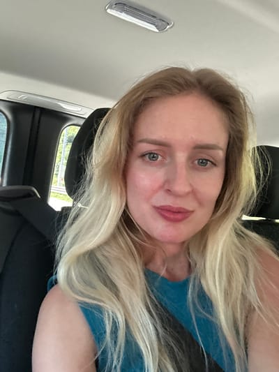 A woman with blonde hair and blue eyes is sitting in the passenger seat of a car, likely in Golling an der Salzach, Austria, as evidenced by the backdrop featuring greenery and what appears to be a roadway. She is wearing a teal-colored top. The image captures her from the chest up, with the car's interior visible in the background, including a seatbelt and the ceiling light. Her expression is relaxed, and she is looking directly at the camera.