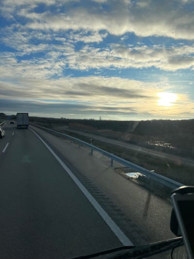The image depicts a highway scene at what appears to be either sunrise or sunset, believed to be in the vicinity of Fraga, Spain. The perspective is from inside a vehicle, looking forward through the windshield, with a portion of a dark object, likely a smartphone, visible in the bottom right foreground.

The sky occupies the upper two-thirds of the frame, featuring a clear blue expanse dotted with numerous white, stratified clouds arranged in horizontal bands across the upper portion. Towards the right horizon, a bright, diffused yellow and white sun is visible, partially obscured by clouds, casting a strong glare. The horizon line itself is low, marked by a distant, flat landscape.

The two-lane asphalt highway dominates the lower left and central sections, with vehicles traveling away from the viewer. A solid white line marks the right edge of the immediate lane. To the right of this line, a rumble strip is present on the paved shoulder, which appears wet in places. A silver metallic guardrail runs along the right side of the highway, extending into the mid-ground. Behind the guardrail, a puddle of water is visible on the paved ground.

In the distance on the highway, a white truck is observed in the right lane, heading away, with other faint vehicle silhouettes visible further ahead in the left lane. Beyond the guardrail and shoulder, the environment transitions to an undeveloped field or embankment with sparse, dark vegetation and dry grasses. Further into the distance on the right, a small, reflective body of water is discernible, mirroring the sky. On the far horizon, a series of dark, indistinct building silhouettes or structures, including a slender spire-like object, suggest a distant settlement or industrial area.