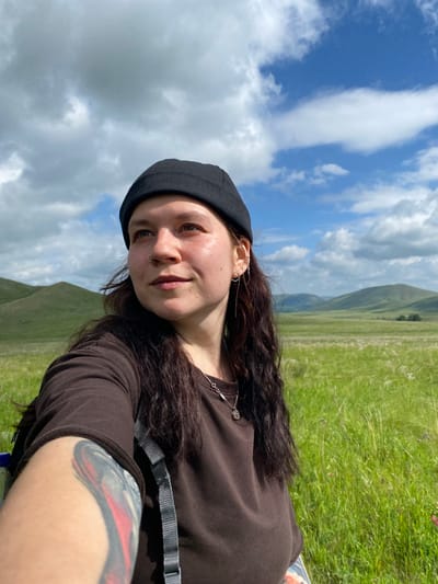 A woman wearing a black beanie and brown t-shirt stands in a grassy field with rolling hills and a cloudy blue sky in the background. She has dark, curly hair, a nose ring, and a tattoo on her arm. The image appears to be a selfie taken outdoors. The location is the city of Andreevka, Russia.