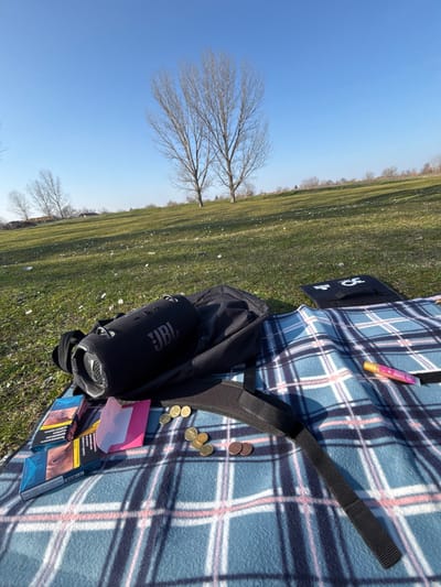 The scene depicts an outdoor leisure setting on a sunny day in the city of Beli Brod, Bulgaria.

The foreground is dominated by a blue and white plaid fleece blanket spread on green grass. On the blanket, a black backpack with a visible shoulder strap rests. Inside or on top of the backpack is a cylindrical black JBL portable speaker, identifiable by its logo. To the left of the backpack, two blue cigarette packs are positioned, one appearing to be open. Scattered nearby are approximately 12-15 metallic coins of varying sizes and colors, consistent in appearance with European small denominations (e.g., Euro cents or Bulgarian stotinki). A small, folded pink paper or wrapper is also present near the cigarette packs. To the right, on the blanket, a slender pink and white cylindrical object, possibly a flavored nicotine pouch or disposable vape, is visible. Further to the right, a flat, rectangular black object, possibly an electronic device or cover, displays a partially visible white logo resembling "3C" or "SC".

The environment consists of a gently sloping grassy hill under a clear, bright blue sky. The grass is green with sparse light-colored debris. In the mid-ground and background, several deciduous trees with bare branches are visible, suggesting late winter or early spring conditions. No individuals are present in the image, but the arrangement of objects suggests recent human activity.