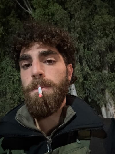 A male individual is depicted in a close-up, chest-level portrait, set against a dark, blurred background of dense green foliage. He has dark, curly hair, a full beard, and a mustache, with brown eyes. He is wearing a dark-colored jacket featuring a prominent silver zipper, layered over an olive-green garment that includes a visible shoulder strap. A lit cigarette, displaying an orange glow at its tip, is held between his lips. His gaze is directed towards the viewer. The environment suggests an outdoor setting at night. The specific location within the Brener Regional Council, Israel, cannot be identified from the visual data present in the image.
