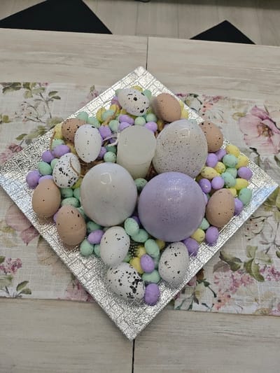 A decorative table centerpiece is captured from an elevated perspective, situated on a table in Opaka, Bulgaria. The central item is a square, silver-colored tray with a textured, possibly hammered or dimpled, surface. The tray is positioned diagonally and contains an assortment of decorative objects. These include numerous small, oval-shaped items, approximately 1-2 cm in length, colored light green, light purple, and yellow, serving as filler material. Interspersed are approximately twelve smaller artificial eggs, measuring 3-4 cm, some plain light beige and others white with black speckles. Five larger artificial eggs are also present, ranging from 5-8 cm in length; these include two smooth white eggs, one white egg with fine black speckles, and one purple egg covered in glitter. A single white, cylindrical object, approximately 6-8 cm tall, consistent with a pillar candle, is centrally located within the arrangement. The tray rests on a textile table runner featuring a light background with a pattern of pink and purple flowers and green leaves. The table runner is placed on a light-colored wooden table, which displays a discernible seam suggesting two joined planks. A dark, angular object is partially visible in the upper background. No individuals are present in the scene.