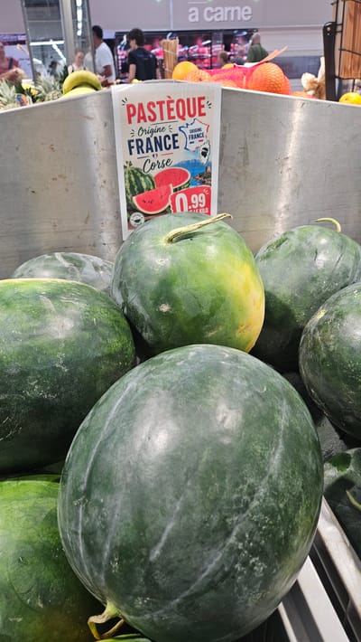 The image shows a close-up view of watermelons for sale in a market or grocery store. A sign in French is prominently displayed, advertising "PASTÈQUE" (watermelon) from "ORIGINE FRANCE Corse." The sign also indicates a price of ".99" per kilogram. Several large, dark green watermelons are visible, with some showing a slight yellowing near the stem. In the background, out of focus, are other fruits like bananas and pineapples, as well as people shopping. The setting appears to be an indoor produce section. The location in Corbara, France is not explicitly visible in the image itself, but the sign suggests a French origin for the watermelons.
