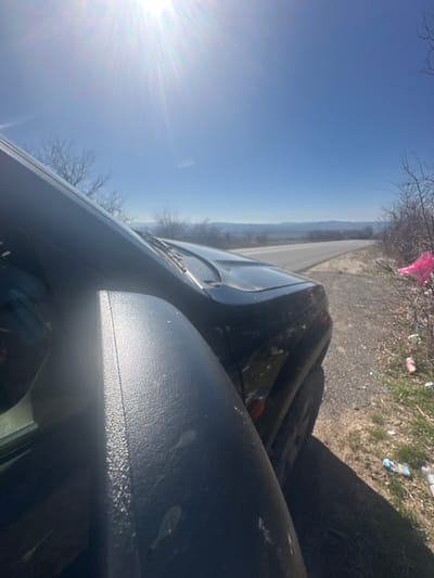 The scene is set on a rural road adjacent to the city of Bogyovtsi, Bulgaria, under a clear, bright blue sky with a high sun causing a significant lens flare in the upper left. A dark-colored passenger vehicle, consistent with an SUV, is stationary on the unpaved right shoulder of a two-lane asphalt road that curves into the distant background. The visible portion of the vehicle includes its front right quadrant, encompassing the hood, a section of the windshield equipped with wipers, and the front right fender. The textured black right side mirror occupies a substantial portion of the immediate foreground, exhibiting visible surface dust. To the right of the vehicle, the roadside features sparse, dry vegetation composed of bushes and bare branches. This area is characterized by substantial litter, including a bright pink plastic bag caught within a bush and several discarded plastic bottles and cans scattered across the ground. The background reveals rolling hills or fields under the clear sky. No individuals are present, and no overt actions or interactions are depicted.