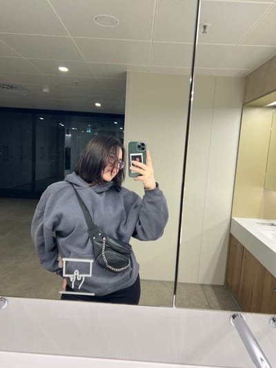A person is reflected in a large mirror, capturing a mirror selfie in a modern, well-lit restroom located in Fischamend, Austria. The individual, with short dark hair and clear-framed glasses, wears an oversized dark grey hoodie and a black fanny pack with a silver chain across their body. They hold a smartphone with a green case and a small square photo attached to its back in their right hand. The reflection reveals a clean environment with light-colored walls and ceiling tiles featuring recessed circular lighting. To the right, two white sinks with chrome faucets are visible above light wood-toned cabinetry. Through a glass partition reflected behind the subject, a dimly lit hallway or interior space with distant lights is discernible. A white graphic, possibly a handwashing icon, is partially visible on the mirror in the lower left foreground.
