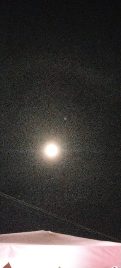 The image depicts a night scene with a prominent, brightly lit full moon in the center of the frame. The moon is surrounded by a halo of light, suggesting it is either very bright or the photograph has been overexposed in that area. The sky is dark and appears to be slightly grainy, possibly due to low light conditions or the camera's sensor noise. Two faint, thin lines, likely power lines or wires, are visible traversing the frame horizontally in the lower half. Below the sky, there is a blurred, light-colored object, possibly a structure or a tent, with some indistinct shapes suggesting it might be a rooftop or a large fabric covering. The overall setting appears to be outdoors, at night, likely in an open area given the view of the sky. There is no visible text in the image. The atmosphere is serene, illuminated by the celestial body.