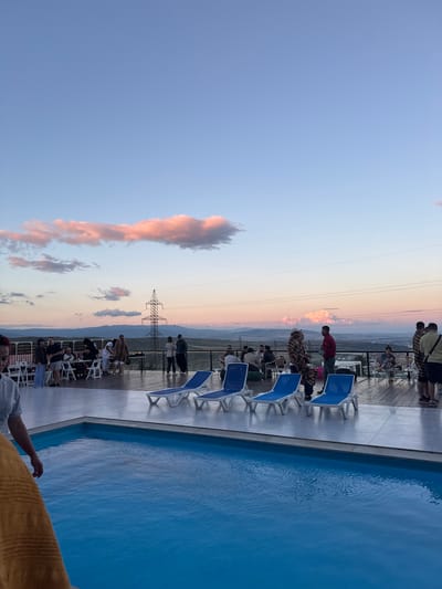 The image depicts an outdoor gathering at dusk, likely in Mukhatskaro, Georgia, with a swimming pool in the foreground. The sky is painted with hues of blue, pink, and orange, indicating a sunset. A group of people are casually dressed and mingling on a raised wooden deck adjacent to the pool. Some are seated at tables, while others stand and converse.
Several blue lounge chairs are neatly arranged near the pool's edge. In the background, a prominent power line tower stands against the horizon, with rolling hills and mountains visible beyond. The overall atmosphere suggests a relaxed social event taking place at a scenic location, possibly a resort or a private residence with a view. The lighting conditions are dim, characteristic of twilight.