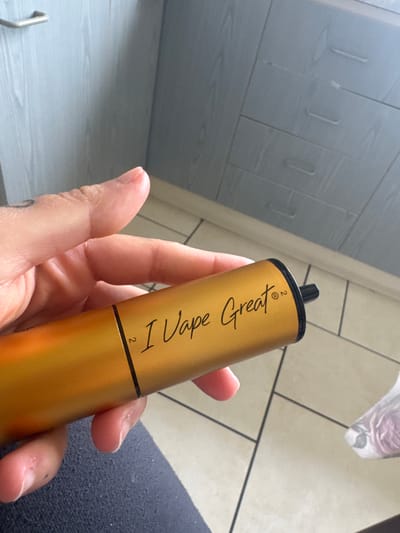 A close-up, eye-level shot shows a person holding a gold-colored disposable vape in their right hand. The vape has black lettering that reads "I Vape Great" with a registered trademark symbol and the number "2" next to it. The background is blurred, showing what appears to be kitchen cabinets with silver handles and tiled flooring. The context of Armagh, United Kingdom is not present in the image.