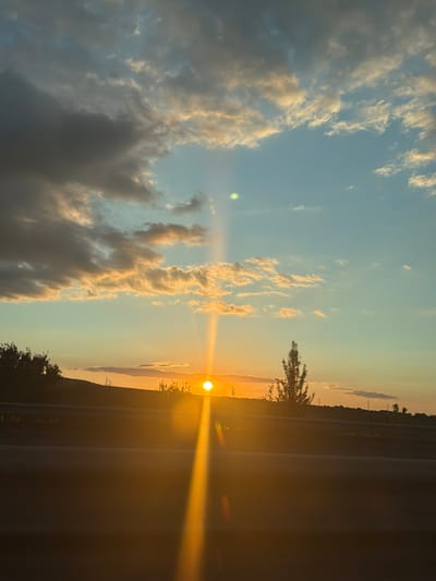 This is a photograph of a sunset over a rural landscape, likely in or near Studena, Bulgaria. The sun is low on the horizon, casting a warm, golden light across the scene. A bright sunbeam extends upwards from the sun, piercing through the scattered clouds. The sky transitions from a clear blue at the top to warmer hues of orange and yellow near the horizon. Darker, more textured clouds are present on the left side of the frame, contrasting with the lighter, wispy clouds illuminated by the setting sun.
In the foreground, a dark, blurry area suggests the perspective is from a moving vehicle, possibly on a road. A horizontal line, appearing to be a barrier or guardrail, runs across the middle ground. Beyond this, the landscape features rolling hills covered with vegetation and trees. A prominent, silhouetted tree stands tall in the middle right of the image. Power lines and poles are faintly visible in the distance along the horizon, indicating some human infrastructure. The overall impression is one of a serene natural setting at dusk.