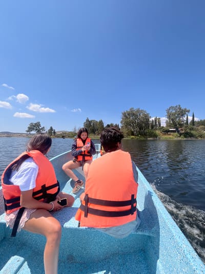 Three individuals, apparently youths, are situated within a light blue boat on the Valsequillo Lake. Each of them is wearing an orange life vest. They are dressed in casual summer clothing. The boat appears to be moving through the water, as evidenced by the wake. The water is dark, and the sky above is bright blue. The vegetation can be seen in the background.